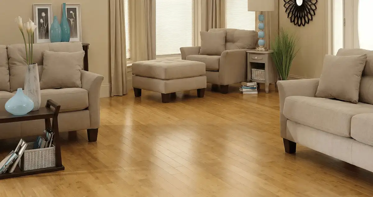 A living room with professionally installed bamboo flooring, beige sofas, a matching ottoman, a side table with a blue lamp, vases, framed pictures, and large windows with blinds and curtains in Chula Vista, CA