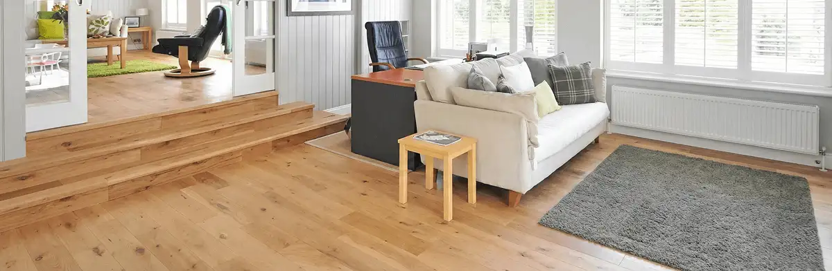 A modern living room with professionally installed laminate flooring, a beige sofa, a small side table, a gray rug, and large windows with white shutters in Chula Vista, CA